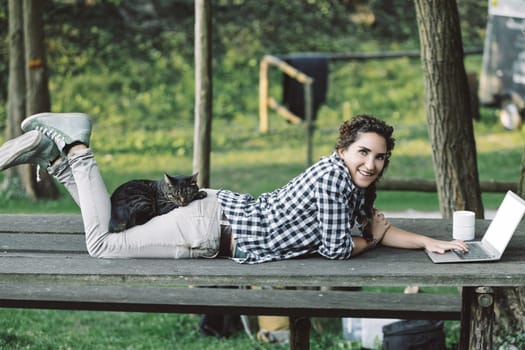 Young woman lies on a picnic table, working on her laptop with her cat sleeping on her back, enjoying the fresh air and sunshine in a peaceful outdoor setting