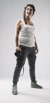 Confident young female photographer looking up and holding a professional camera, she is wearing casual clothing and posing in a studio with a white background