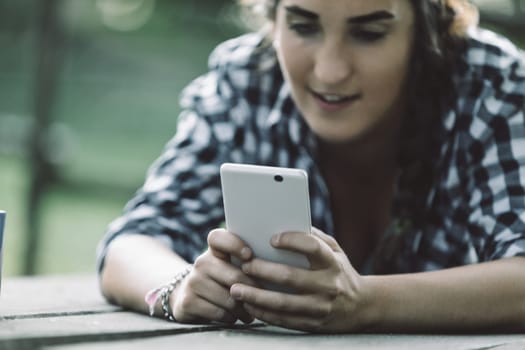 Young woman lying on a wooden table outdoors, using a tablet while enjoying a peaceful moment in nature. Surrounded by greenery, she connects with the digital world in comfort