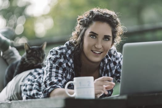 Young woman is lying on her stomach on a wooden table outdoors, working on her laptop with her cat lying on her back and a mug beside her