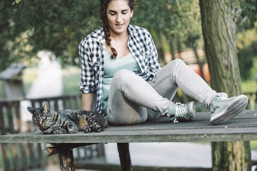 Young woman with braided hair, wearing a plaid shirt and jeans, sits on a wooden table outdoors with her tabby cat resting beside her, enjoying a peaceful moment in nature