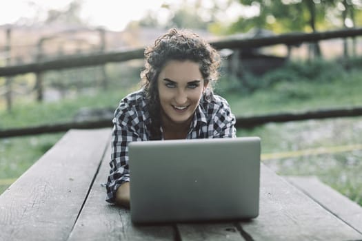 Smiling young woman lying on a wooden picnic table in a rural setting, using her laptop for online learning, enjoying the flexibility and convenience of remote education
