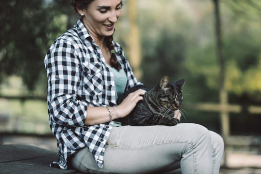 Young woman sitting on a bench outdoors in a park is petting a tabby cat lying on her lap, enjoying a peaceful moment of connection with nature and her feline companion