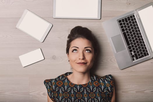 Young woman lies on a wooden floor surrounded by digital devices, lost in thought and gazing up, capturing the essence of the modern digital age