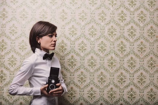 Young female photographer holding a vintage twin-lens reflex camera while posing against a patterned wallpaper, creating a nostalgic and artistic atmosphere