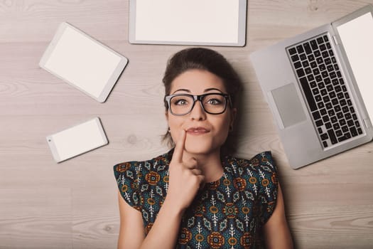 Young woman with eyeglasses lying on the floor surrounded by a laptop, tablet and smartphone, all with blank white screen, touching her chin with her finger and thinking