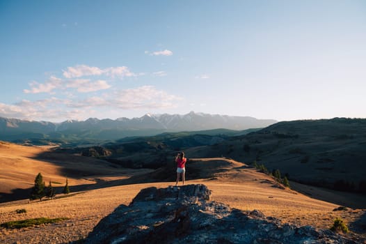 A woman in casual clothing taking photo on her smartphone on a rocky peak, gazing at scenic mountains and a vast valley under golden sunlight.