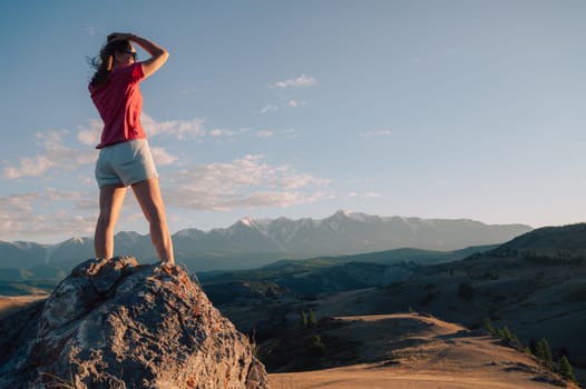 A woman in casual clothing stands on a rocky peak, gazing at scenic mountains and a vast valley under golden sunlight.