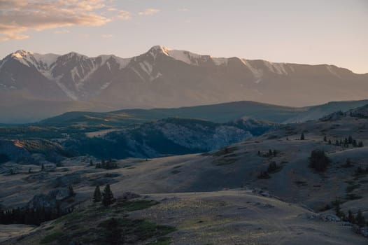 A breathtaking sunset over a rugged mountain landscape, casting warm golden light across hills, trees, and rocky terrain. Altai Mountains