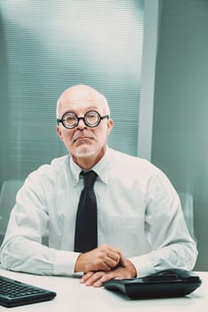 Nerdy businessman with thick glasses sitting at his desk, gazing seriously at the camera while surrounded by office essentials, conveying a professional and focused demeanor