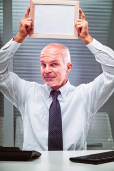Mature businessman sitting at his desk, holding a blank frame above his head while looking at the camera with a humorous expression, inviting creativity and imagination