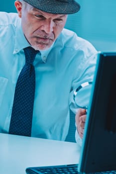 Businessman wearing a hat and tie is carefully examining his computer screen with a magnifying glass, demonstrating deep concentration and attention to detail