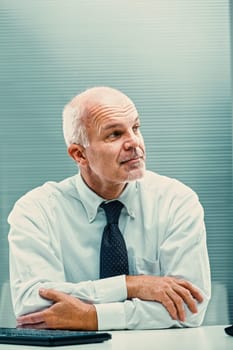 Thoughtful mature businessman sitting at his desk with arms crossed, looking away in contemplation, perhaps considering a new business strategy or reflecting on a challenging decision