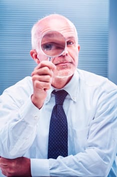 Mature businessman holding a magnifying glass over his eye, looking closely at something, with arms crossed, wearing a light blue dress shirt and tie, in an office setting
