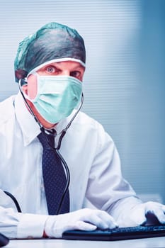 Doctor wearing a surgical mask, hairnet, and gloves, typing on a keyboard while making eye contact with the camera, showcasing a stethoscope around his neck in a clinical setting