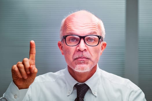 Mature businessman raising his index finger in a warning gesture, exuding seriousness while wearing a crisp white shirt and tie in a professional office setting