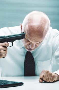 Senior businessman pointing a gun to his head while sitting at his desk in the office, he is stressed and desperate because of overwork and financial problems
