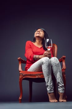 Young woman laughing while holding a wineglass, sitting barefoot on a vintage armchair and gazing upwards, embodying joy and carefree relaxation in a stylish indoor setting