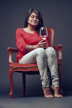 Young woman with long dark hair, wearing a red shirt and patterned pants, sits barefoot on an antique wooden chair, holding a glass of white wine and looking at it with a thoughtful expression