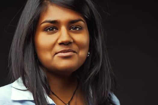 Portrait of a young indian woman with long black hair and brown eyes, wearing a light blue shirt, looking directly at the camera with a thoughtful expression on a dark background