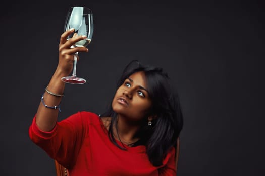 Young woman with long black hair wearing a red shirt holds a glass of white wine up to the light and examines it against a dark studio background