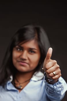 Young indian businesswoman expressing mixed feelings while showing a thumbs up sign of approval, slightly frowning yet radiating confidence and determination in a professional setting