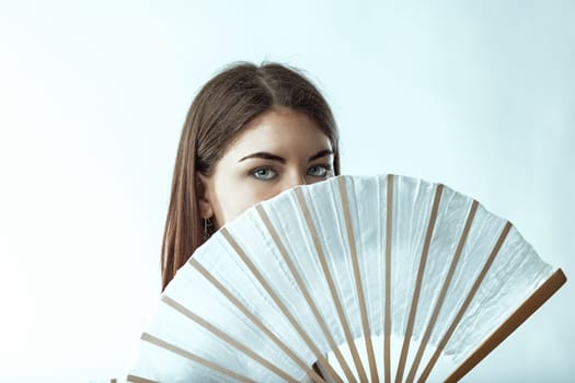 Studio portrait of a mysterious young woman with blue eyes partially hiding her face behind a white hand fan, creating an intriguing and sensual atmosphere