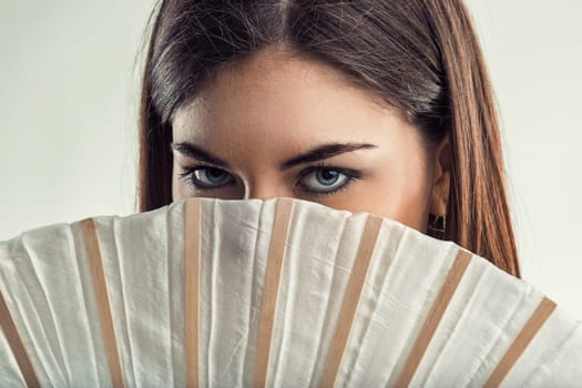 Captivating close-up portrait of a young woman with striking blue eyes peering over a white fan, creating an aura of mystery and intrigue