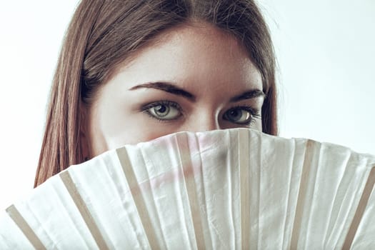 Close-up portrait of a young woman with striking blue eyes gazing over a delicate white hand fan, exuding an enchanting air of mystery and playful intrigue