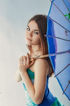 Beautiful young woman holding a decorated blue parasol on her shoulder, smiling and looking directly at the camera, exuding charm and elegance against a white background