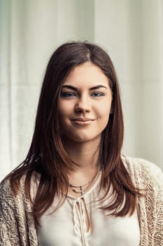 Portrait featuring a beautiful young woman with long brown hair and captivating blue eyes, smiling confidently at the camera, radiating happiness and positivity in a casual white shirt and cardigan