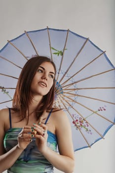 Young woman holding a traditional light blue parasol with flower decorations, looking away from the camera with a thoughtful expression, posing for a fashion or beauty portrait