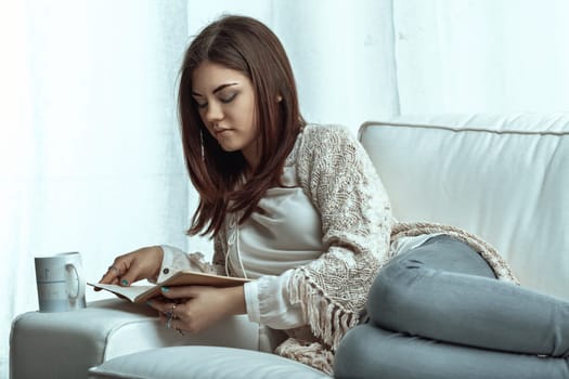Young woman lounging on a white sofa in front of a window, wearing casual clothes, deeply engrossed in a book while savoring a warm cup of coffee, enjoying a peaceful moment