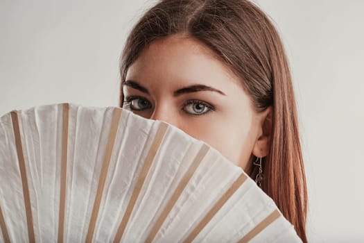 Studio shot of a young woman with brown hair and light eyes partially covering her face with a white and beige hand fan, creating a sense of mystery and intrigue