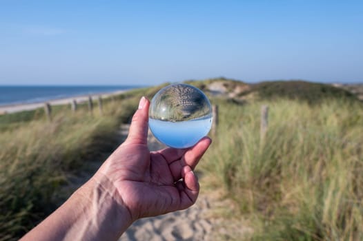 A human hand holds a glass ball, in the ball a sand dune, a footpath, blue sky and the sea are reflected