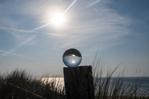 Glass ball lies on a wooden post, beach grass in the background, the sea, the sun and the blue sky are reflected in the ball