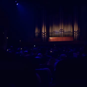 A large illuminated pipe organ in a dark concert hall with a seated audience, creating a dramatic and atmospheric scene.
