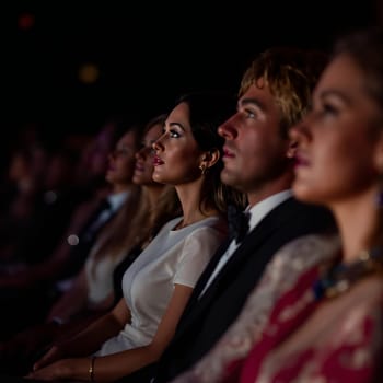 A group of elegantly dressed people attentively watching a performance in a dimly lit theater.