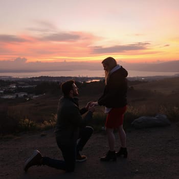 A man proposing to a woman at sunset on a hill, with a breathtaking view of the city and landscape in the background.