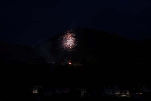 A firework rocket in the sky on a mountain at night, a few illuminated houses in the foreground