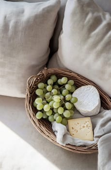 A woven basket holds green grapes and two types of cheese resting on a soft fabric. Sunlight filters softly over the scene.