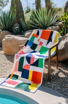 A vibrant towel featuring geometric patterns is elegantly draped over a wooden chair near a sunny poolside, surrounded by lush greenery.