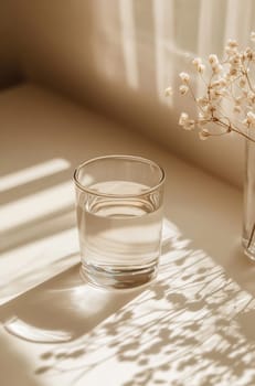 A clear glass filled with water sits on a table next to a vase of small white flowers, basking in soft sunlight.