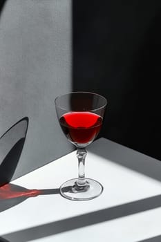 A clear glass with a red liquid sits on a white table, illuminated by natural light creating striking shadows.