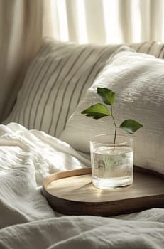 A glass of water with a leafy sprig sits on a wooden tray beside soft, inviting linens in a sunlight-filled room.