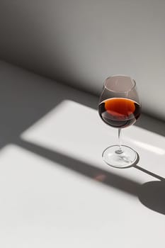 A wine glass filled with dark, rich liquid sits on a table, illuminated by soft natural light casting a long shadow.