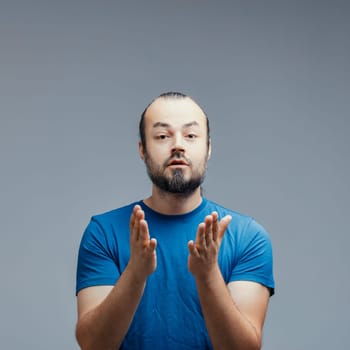 Caucasian man with beard and ponytail, wearing blue t-shirt, looking surprised while making welcome and agreement gestures with hands, isolated on gray background