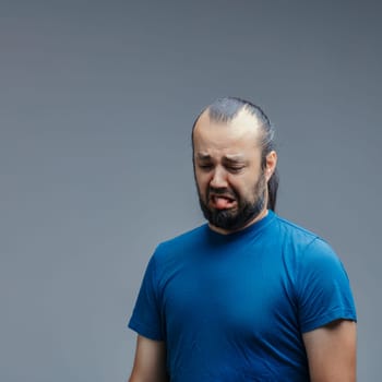 Mid adult man with a beard and ponytail, wearing a blue t-shirt, sticking out his tongue in disgust while looking down against a gray background, expressing strong aversion
