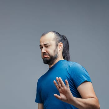 Man with long dark hair in a ponytail, wearing a blue t-shirt, making a disgusted face and raising his hand in rejection against a gray backdrop