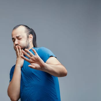 Young adult man with beard and long hair tied in a ponytail wearing a blue t-shirt is expressing disgust and covering his mouth with his hand while also pushing away with the other hand
